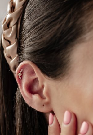 Goldsternförmige Ohrringe mit klaren Akzenten, die im Ohr getragen werden. Das Haar ist dunkel und glatt, mit einem hellbeigen geflochtenen Haarband. Die Nägel sind hellrosa.