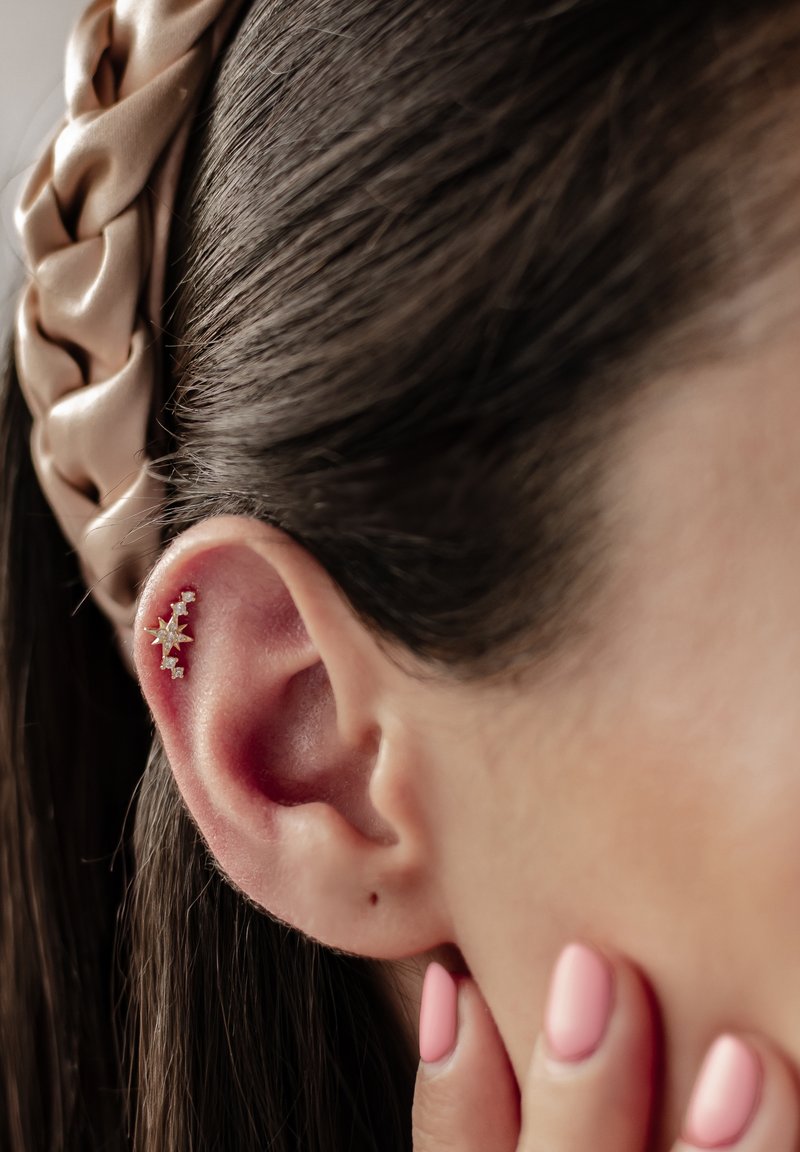 Boucle d'oreille en forme d'étoile dorée avec des accents transparents, présentée dans l'oreille. Les cheveux sont noirs et lisses, avec un bandeau tressé beige clair. Les ongles sont rose clair.