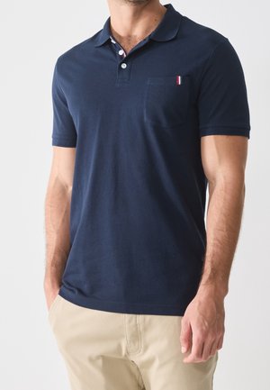 Homme portant un polo bleu marine à manches courtes avec une poche poitrine et un pantalon beige, debout devant un fond clair uni.
