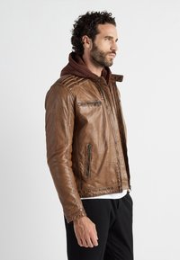 Ruskea nahkatakki, jossa on muotoiltu malli, vetoketjulliset taskut ja päällitikkaukset olkapäillä. Alla on ruskea huppari.