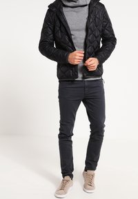 Svart quiltad jacka med huva, bärs över en grå tröja. Matchar med marinblå slim-fit byxor och beige sneakers. Slät textur, praktisk design.