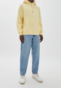 Žlutá mikina s kapucí a klokaní kapsou, nošená s volnými světle modrými džínami a bílými teniskami na jednoduchém pozadí.
