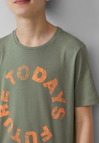 Olijfgroen katoenen T-shirt met een cirkelvormige graphic en gestructureerde oranje letters die "DE TOEKOMST VANDAAG" lezen. Ronde hals en korte mouwen.
