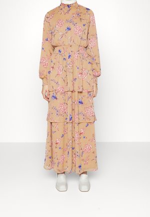 Langes Blumenkleid in Beige mit rosa und blauen Blumen. Es verfügt über gestufte Lagen und einen hohen Kragen, kombiniert mit weißem Schuhwerk.