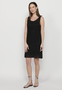 Abito nero all'uncinetto con design senza maniche, motivo a strati e orlo scallop. Abbinato a sandali neri con cinturini per un look completo.