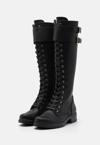 Black knee-high boots made of smooth leather, featuring a lace-up front, gold-tone eyelets, and two decorative straps at the top.