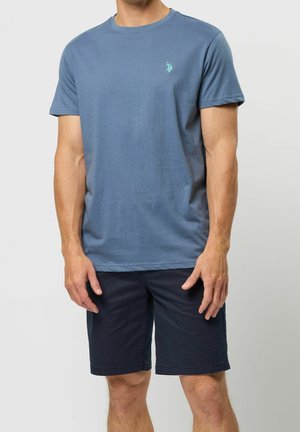Uomo che indossa una t-shirt blu a girocollo con piccolo logo sul petto e pantaloncini blu scuro fino al ginocchio, in piedi con le braccia rilassate.