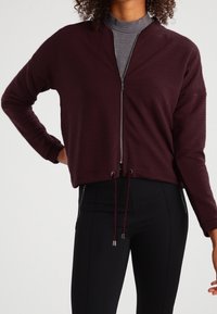 Burgundin värinen lyhyempi takki, jossa on tekstuuri, vetoketjukiinnitys ja narut. Wear up gray turtleneckin päällä, yhdistettynä mustiin leggingsiin.