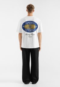 Hvid bomuld t-shirt med et blåt og guld grafisk design på ryggen. Teksten "SAPIENTA ET VERITAS" og "Heritage Center" er fremhævet.
