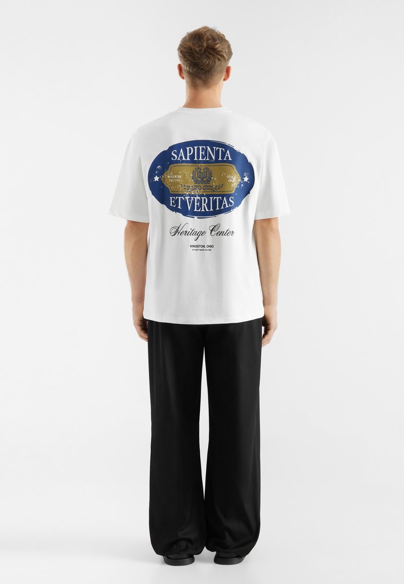 Hvid bomuld t-shirt med et blåt og guld grafisk design på ryggen. Teksten "SAPIENTA ET VERITAS" og "Heritage Center" er fremhævet.