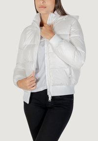 Giacca a vento bianca con chiusura a zip, polsini a coste e cappuccio. Texture liscia, design trapuntato con cuciture visibili, indossata sopra una maglietta bianca.