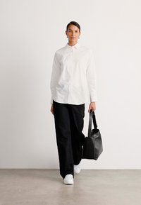 Camicia bianca a maniche lunghe con bottoni e motivi sottili, abbinata a pantaloni neri a gamba larga e sneakers bianche. Tiene in mano una borsa tote nera.