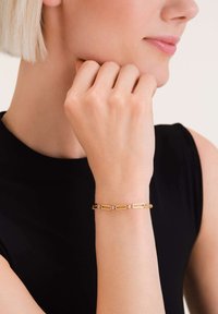 Pulsera de oro con formas de eslabones alargados y pequeños acentos decorativos. Acabado suave y textura pulida, llevada en una muñeca.