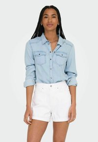 Camisa de mezclilla azul claro con dos bolsillos en el pecho, botones en la parte delantera y mangas enrolladas; combinada con shorts de mezclilla blancos con dobladillo. 