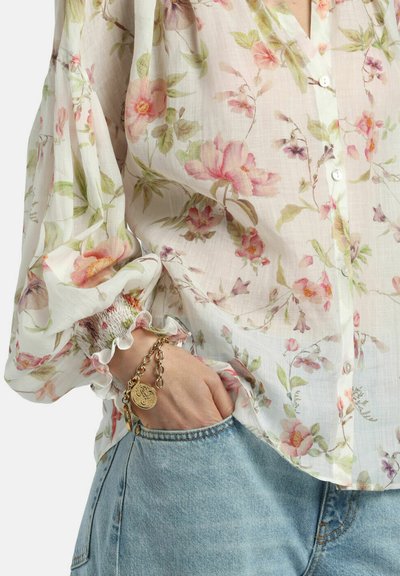 Kvinde iført gennemsigtig blomstret bluse med puffede ærmer og blå denimjeans, hånd i lommen, guldbelagt charm-armbånd på håndleddet.