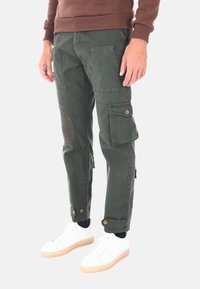 Pantaloni cargo verdi realizzati in cotone, con tasche laterali, cuciture rosse sulle ginocchia e una vestibilità affusolata. Indossati con scarpe da ginnastica bianche.