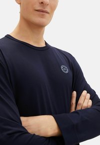 Marineblauw langemouwen shirt van zachte stof, met een ronde hals en een klein logo op de linkerborst. Gladde textuur, aansluitend ontwerp.