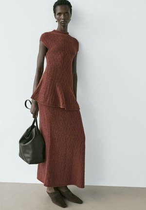 Modella che indossa un abito di due pezzi di colore ruggine con texture, top svasato e gonna lunga, con in mano una borsa nera in pelle e scarpe scamosciate marrone scuro con punta appuntita.