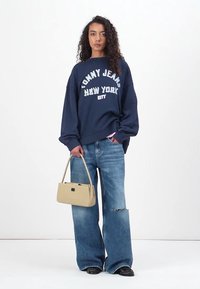 Felpa oversize blu navy con stampa "Tommy Jeans New York", abbinata a jeans a gamba larga blu chiaro, borsa beige e scarpe nere.