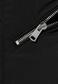 Detail čiernej bundy so zipsom, ktorý má kovový zips s obdĺžnikovým ťahátkom na hladkom, matnom povrchu.