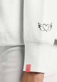 Witte sweatshirt met een hart met vleugels ontwerp op de mouw en een klein roze label met een grafisch accent. Zachte textuur, casual pasvorm.