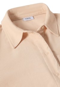 Cuello de camisa de pana beige y los botones superiores visibles, con la etiqueta de la marca CECIL en el cuello.