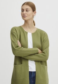 Cardigan vert olive avec un tricot texturé, ouverture sur le devant, deux poches avant et poignets côtelés. Porté sur une chemise blanche avec un col rond.