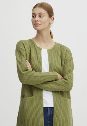 Cardigan vert olive avec un tricot texturé, ouverture sur le devant, deux poches avant et poignets côtelés. Porté sur une chemise blanche avec un col rond.