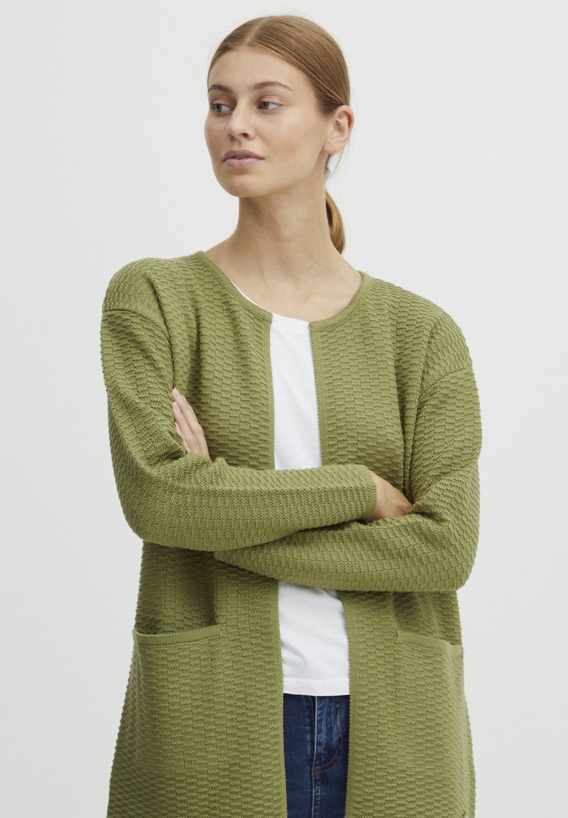 Cardigan vert olive avec un tricot texturé, ouverture sur le devant, deux poches avant et poignets côtelés. Porté sur une chemise blanche avec un col rond.