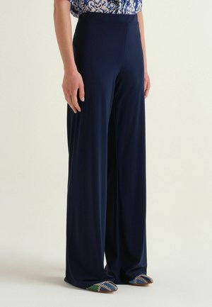 Persona che indossa pantaloni blu navy a vita alta e a gamba larga abbinati a una maglietta fantasia e scarpe multicolori a punta, in piedi davanti a uno sfondo semplice.