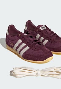 Ζευγάρι μπορντό αθλητικά παπούτσια Adidas Japan με λευκές ρίγες και λαστιχένιες σόλες, συνοδευόμενα από επιπλέον λευκά κορδόνια σε λευκό φόντο.