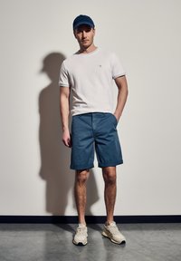 Bílý tričko s krátkým rukávem a černými detaily, modré šortky a béžové tenisky. Model má také na sobě modrou čepici a stojí před světlou zdí.