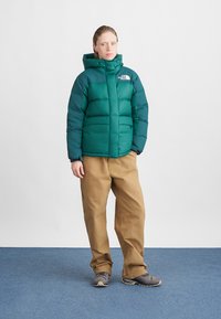 Green puffer jacket with a hood, featuring a zip closure and a logo on the chest, paired with beige pants and gray shoes.