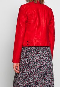 Veste en cuir rouge avec des accents matelassés aux épaules et des détails à pression au bas, portée sur une robe florale navy. Texture lisse, design cropped.