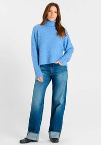 Vrouw in een blauwe coltrui, met wijde, opgerolde jeans en zwarte schoenen, staande met één hand in de zak.