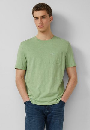 Jeune homme aux cheveux courts et bouclés portant un t-shirt vert clair à manches courtes avec une poche poitrine et un jean bleu foncé, se tenant contre un mur gris.