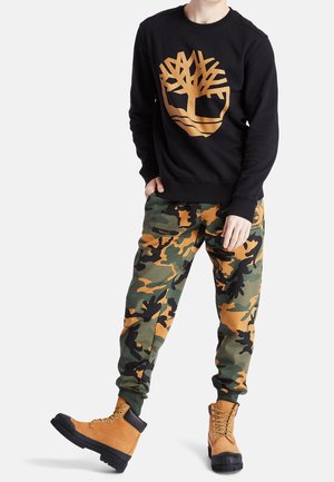 Persoon draagt een zwart sweatshirt met een oranje boomlogo, groene en oranje camouflagebroek en beige veterlaarzen met zwarte zolen.