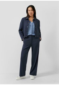 Marineblauw cropped jack met knoopsluiting, gecombineerd met een lichtblauw textuurtopje en marineblauwe wijde pantalon. Model draagt witte sneakers.