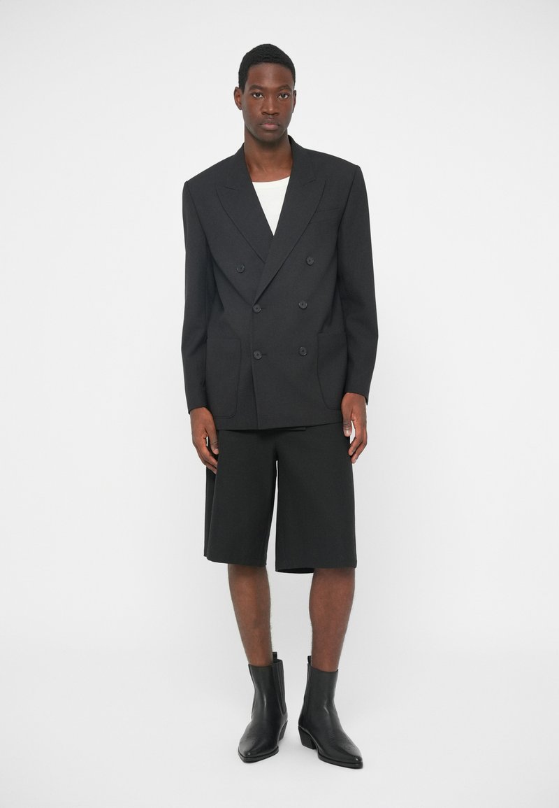 Sort dobbeltradet blazer med revers, parret med sorte skræddersyede shorts og sorte ankelstøvler, sat mod en hvid baggrund.