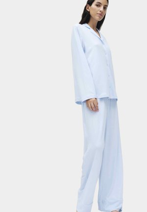 Kvinde stående iført lyseblåt, løstsiddende pyjamassæt med knapper, lange ærmer og vidde bukser mod en ensfarvet baggrund.