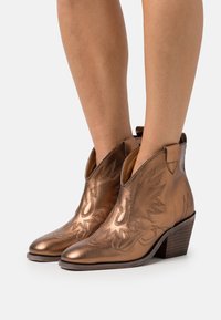 Bottines en cuir métallisé bronze avec des coutures de style western, portées sur des jambes nues, avec un talon empilé et une pointe effilée.