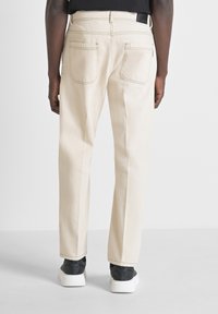 Pantalones de algodón color beige con corte recto, que presentan dos bolsillos traseros y costuras en contraste, llevados con zapatillas oscuras.