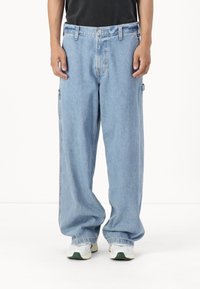 Kraťasy z lehkého modrého denimu s vysokým pasem, zapínáním na knoflík vpředu a bočními kapsami. Nosí se s šedými a zelenými teniskami.