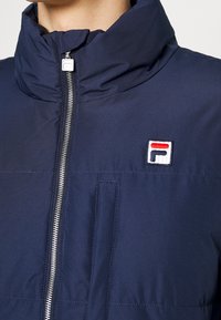 Marinblå zip-up jacka med hög krage, prydd med en Fila-logotyp och framficka, tillverkad av ett mjukt och lätt material.