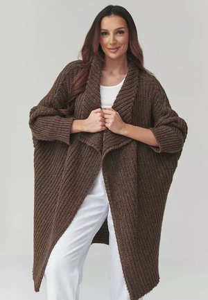 Kobieta ubrana w długi, oversize brązowy sweter z dzianiny noszony na białym topie i białych spodniach, trzymająca sweter zapięty na piersi.