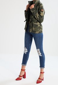 Olivově zelená maskáčová bunda, černý top, modré džínové kalhoty s popraskaným vzhledem a červené lodičky na podpatku s kotníkovými pásky. Poznámka: roztrhané detaily na džínách.