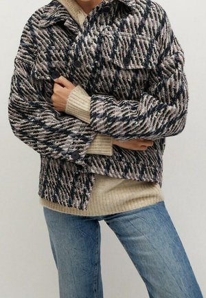Person iført en tekstureret sort, hvid og beige hundetandsjakke over en beige ribbet sweater og blå jeans med armene krydset.