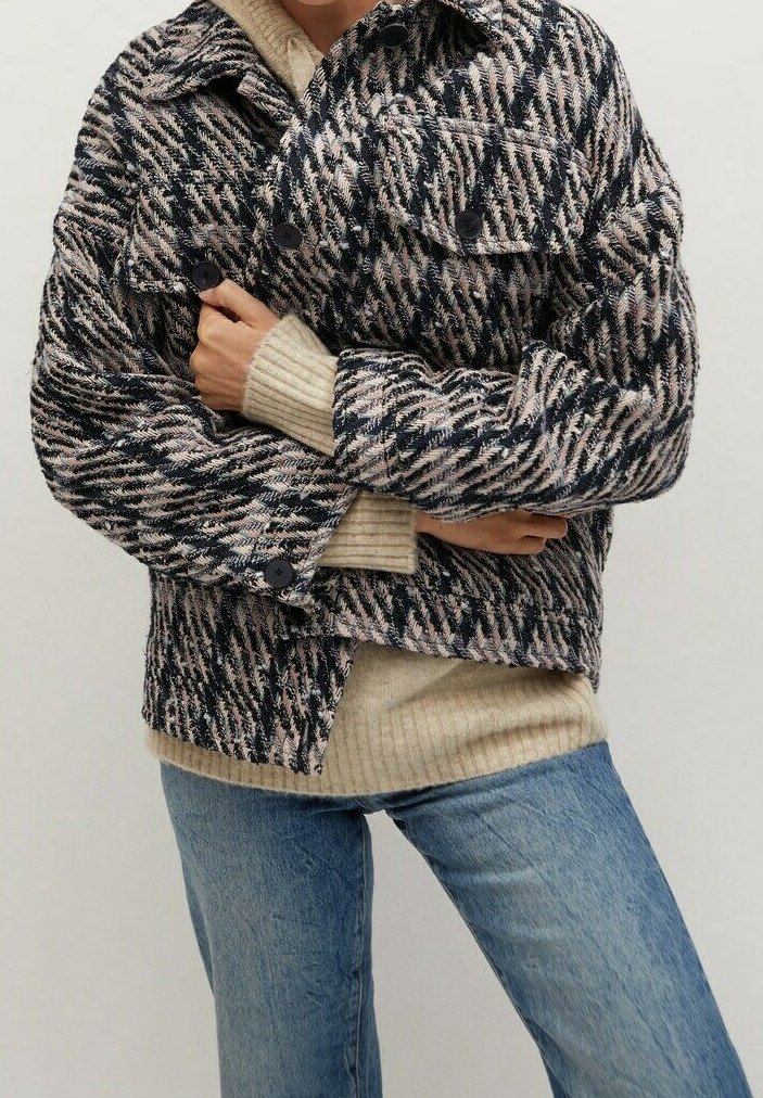 Personne portant une veste pied-de-poule texturée noire, blanche et beige sur un pull côtelé beige et un jean bleu, les bras croisés.