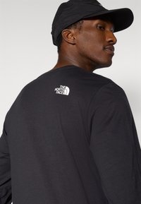 Μαύρο μακρυμάνικο πουκάμισο από μαλακό ύφασμα με στρογγυλή λαιμόκοψη. Διαθέτει λευκό λογότυπο στο πάνω μέρος της πλάτης. Φοράται με μαύρο καπέλο.