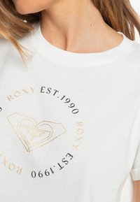 Weißes Baumwoll-T-Shirt mit einem runden Logo-Design, das "Roxy" und "Est. 1990" in Schwarz und Gold zeigt. Angenehme Textur und Rundhalsausschnitt.
